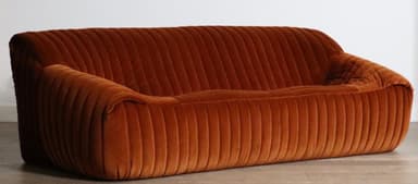 A plush, channel-tufted sofa upholstered entirely in a warm, burnt orange velvet fabric. It features a low-profile design with rounded contours and integrated armrests that seamlessly flow into the backrest and base. The distinctive vertical channel tufting provides a rich texture across all upholstered surfaces. — 3D model