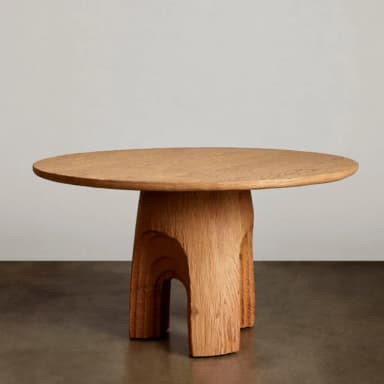 A round wooden coffee table featuring a thick, sculptural pedestal base with deeply carved, layered arch motifs. The solid wood has a visible grain and a textured, rustic finish in a light brown hue. — 3D model