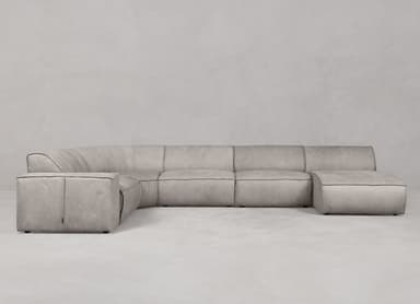 A contemporary modular sectional sofa featuring an L-shaped configuration with a curved corner unit and an integrated ottoman extension. The sofa is upholstered in a light grey or greige fabric with prominent black contrast stitching along all seams, highlighting its clean, architectural lines. It has a low-profile design with wide, flat armrests and seat cushions, suggesting a modern and minimalist aesthetic. — 3D model