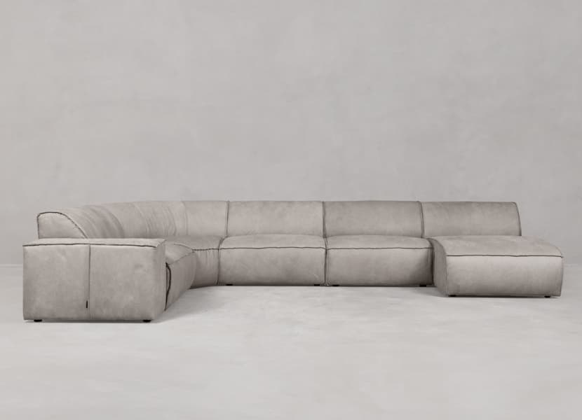A contemporary modular sectional sofa featuring an L-shaped configuration with a curved corner unit and an integrated ottoman extension. The sofa is upholstered in a light grey or greige fabric with prominent black contrast stitching along all seams, highlighting its clean, architectural lines. It has a low-profile design with wide, flat armrests and seat cushions, suggesting a modern and minimalist aesthetic. — 3D model