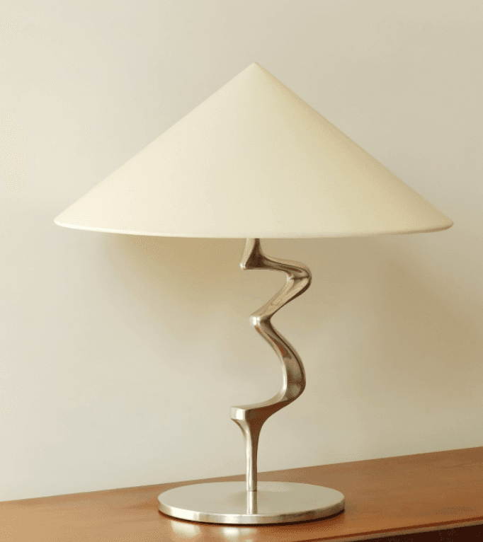 A modern table lamp featuring a distinctive serpentine metal stem, a circular base, and a conical fabric shade. The design emphasizes sculptural form and a polished finish. — 3D model