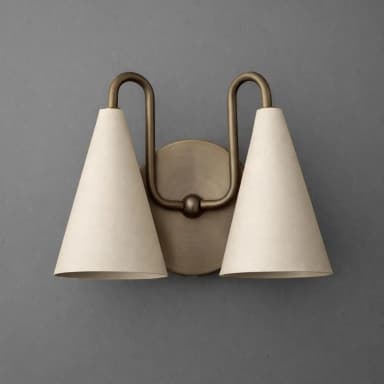 A two-light wall sconce featuring an antique brass finish metal frame and two conical shades. The fixture mounts to the wall via a circular backplate, from which two U-shaped arms extend upwards and curve down to hold the individual lamp shades. The shades are off-white, likely fabric, with a distinct conical shape. — 3D model