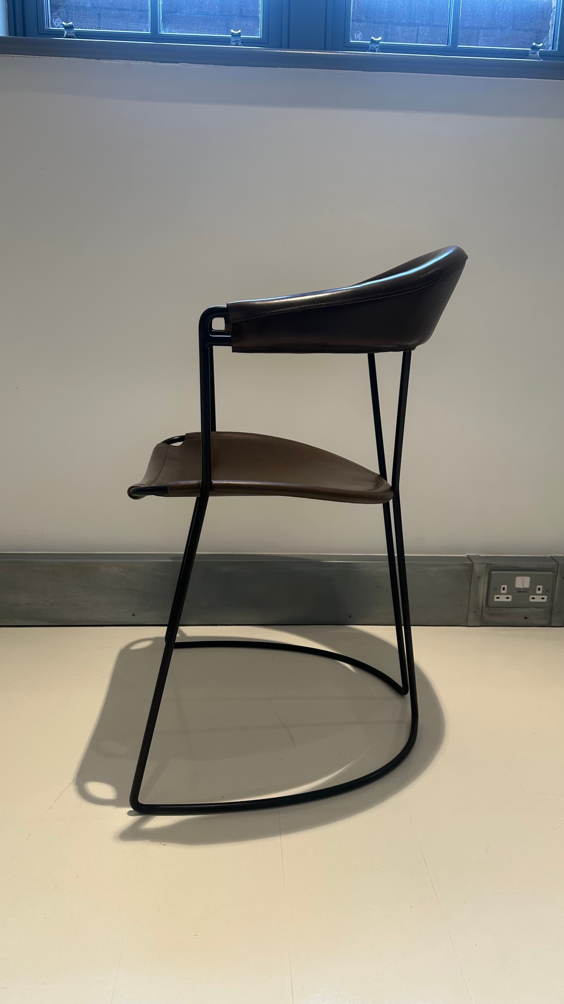 A contemporary armchair with a dark brown leather seat and backrest supported by a slender black metal frame. The frame features a curved base that extends into the legs, providing stability and a dynamic visual element. The seat and backrest are subtly contoured for comfort and appear to be constructed from stitched leather panels. — 3D model