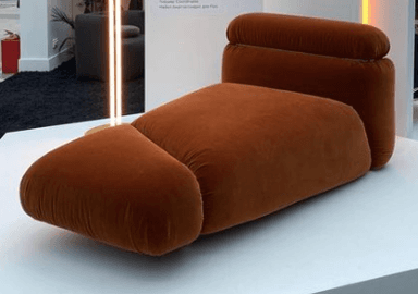 A plush, contemporary chaise lounge upholstered in a warm, rust-colored velvet-like fabric, featuring soft, rounded forms and a distinct extended footrest section. — 3D model