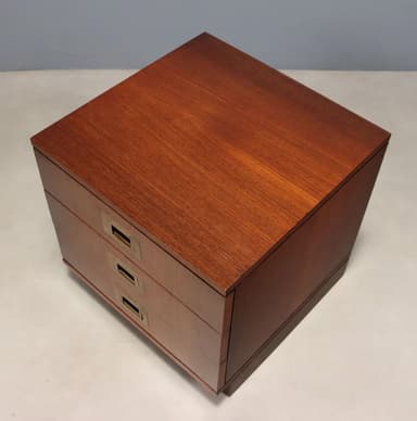 A compact, three-drawer wooden nightstand or side table, characterized by its reddish-brown finish and distinctive brass campaign-style recessed pull handles, supported by a recessed plinth base. — 3D model