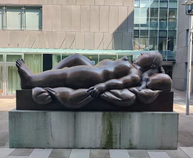 A large bronze sculpture depicting a reclining female figure, situated outdoors on a substantial concrete plinth in front of a modern glass building. The figure has soft, rounded forms and is posed with her head resting on a raised form resembling a pillow, with her body gently draped. — 3D model