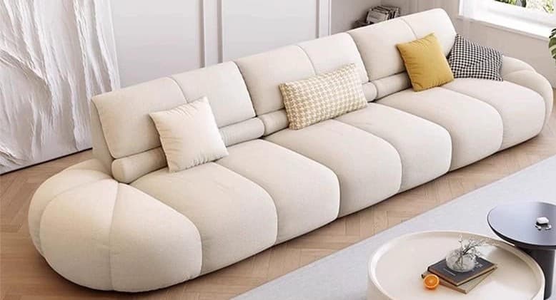 A long, low-profile sofa with a rounded, segmented base and backrest. The sofa features tufted or channel-stitched upholstery. It is accompanied by four throw pillows of varying sizes and patterns: two rectangular pillows with a subtle checkered pattern, one square cushion in a solid yellow, and one square cushion with a black and white gingham pattern. The overall design is contemporary with a soft, inviting aesthetic. — 3D model