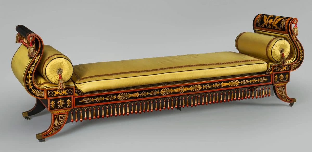 A sophisticated Neoclassical style daybed featuring a long, rectangular cushioned surface upholstered in yellow fabric, flanked by elegant scrolled ends. The wooden frame is lacquered black with intricate gilded and red painted classical motifs, including stylized wings and foliage. Decorative tassels in red and gold adorn the side frame, enhancing its luxurious and historical aesthetic. The daybed stands on short, curved legs. — 3D model