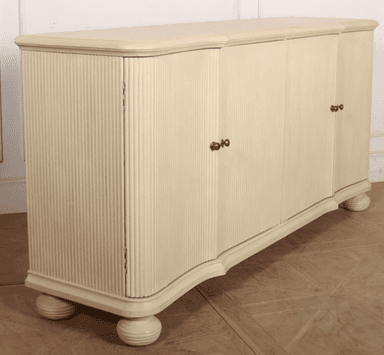 A long, cream-colored sideboard featuring four doors and a subtly curved top and base. The end sections and outer doors are adorned with vertical reeded detailing, while the two central doors are smooth. The piece stands on four spherical, turned feet and is outfitted with small, ornate brass-tone door pulls. — 3D model