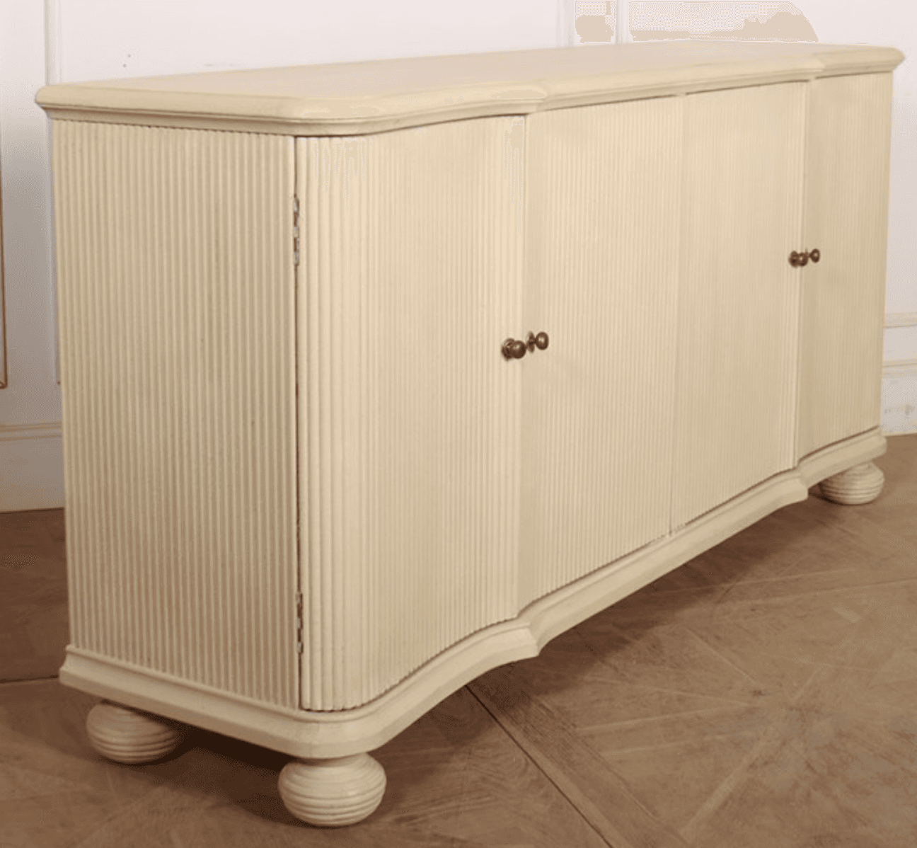 A long, cream-colored sideboard featuring four doors and a subtly curved top and base. The end sections and outer doors are adorned with vertical reeded detailing, while the two central doors are smooth. The piece stands on four spherical, turned feet and is outfitted with small, ornate brass-tone door pulls. — 3D model