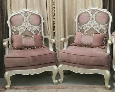 A single upholstered armchair with an ornate carved wooden frame, featuring a high, slightly curved back and padded armrests. The backrest is upholstered in a floral pattern of pink and off-white, while the seat cushion and armrests are solid pink. The frame is finished in a light, possibly cream or silver-gilt tone, with cabriole legs and scrolled details. A small matching bolster pillow with ruffled trim is included. — 3D model
