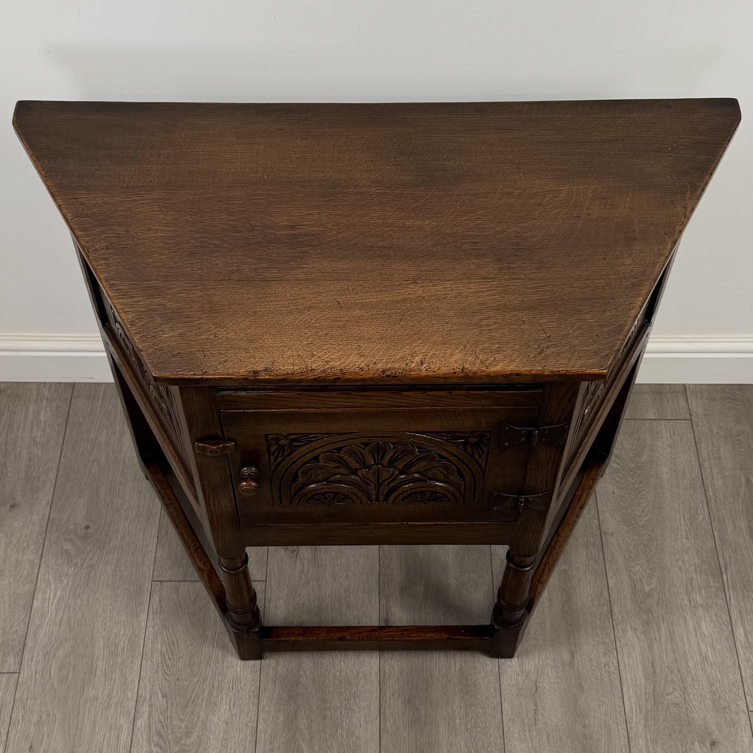 A vintage wooden cabinet with a triangular top. The front features carved decorative elements, a hinged door with a small knob handle, and metal hinges. The cabinet has four legs, two of which are visible, and a stretcher connecting them near the bottom. The wood grain is prominent, and the finish is dark brown. It appears to be a small accent cabinet or a cellarette. — 3D model