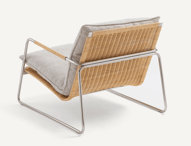 A low-profile modern lounge chair featuring a sleek, polished tubular steel frame. The seat and backrest are constructed from tightly woven natural cane, providing a textured, organic element. Thick, light-colored fabric cushions rest on the cane surfaces for comfort. The integrated armrests are part of the continuous steel frame, with a wrapped detail on the top sections. — 3D model