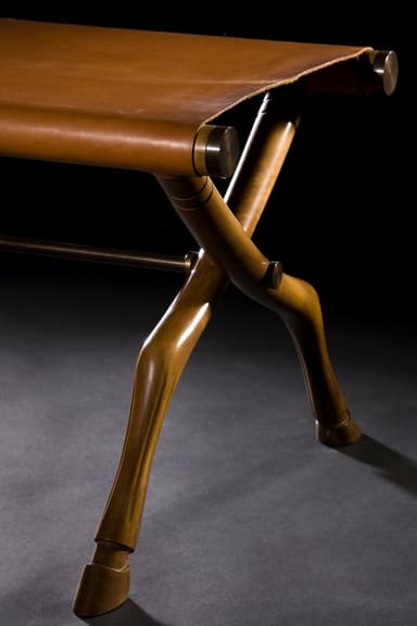 A campaign-style folding stool featuring an X-shaped wooden frame with carved details and a tan leather sling seat. The legs are notably carved to resemble animalistic or hoofed forms, adding an exotic touch. The frame is joined by visible metal hardware at the pivot points and where the leather seat attaches. — 3D model
