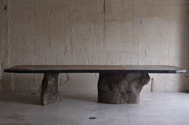 A large dining table with an irregular, organic-shaped top and two substantial, textured bases that resemble natural rock formations or tree trunks. The tabletop appears to be made of multiple panels, possibly wood or stone, with a dark, possibly metal or resin, edge banding. The bases are dark brown and have a rough, sculpted texture, giving the impression of raw, natural elements. — 3D model