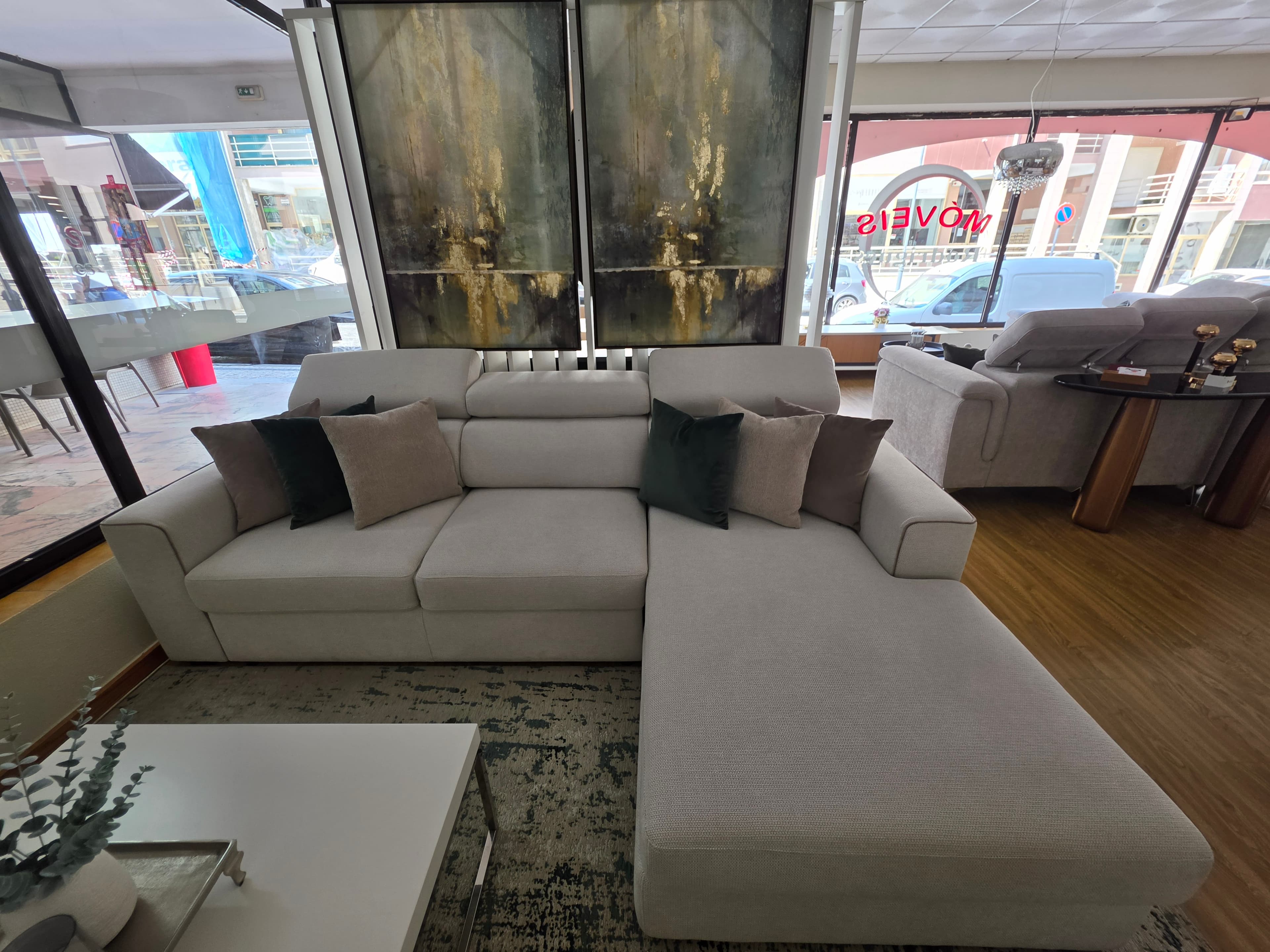 An L-shaped sectional sofa upholstered in a light beige fabric, featuring an integrated chaise lounge on the right side. The sofa is composed of modular sections with clean lines and subtly piped seams. It includes multiple adjustable headrests that can be folded down when not in use. Several accent pillows in varying shades of green and brown are placed on the sofa, adding a touch of color. The base of the sofa is low-profile with dark, discreet feet. — 3D model