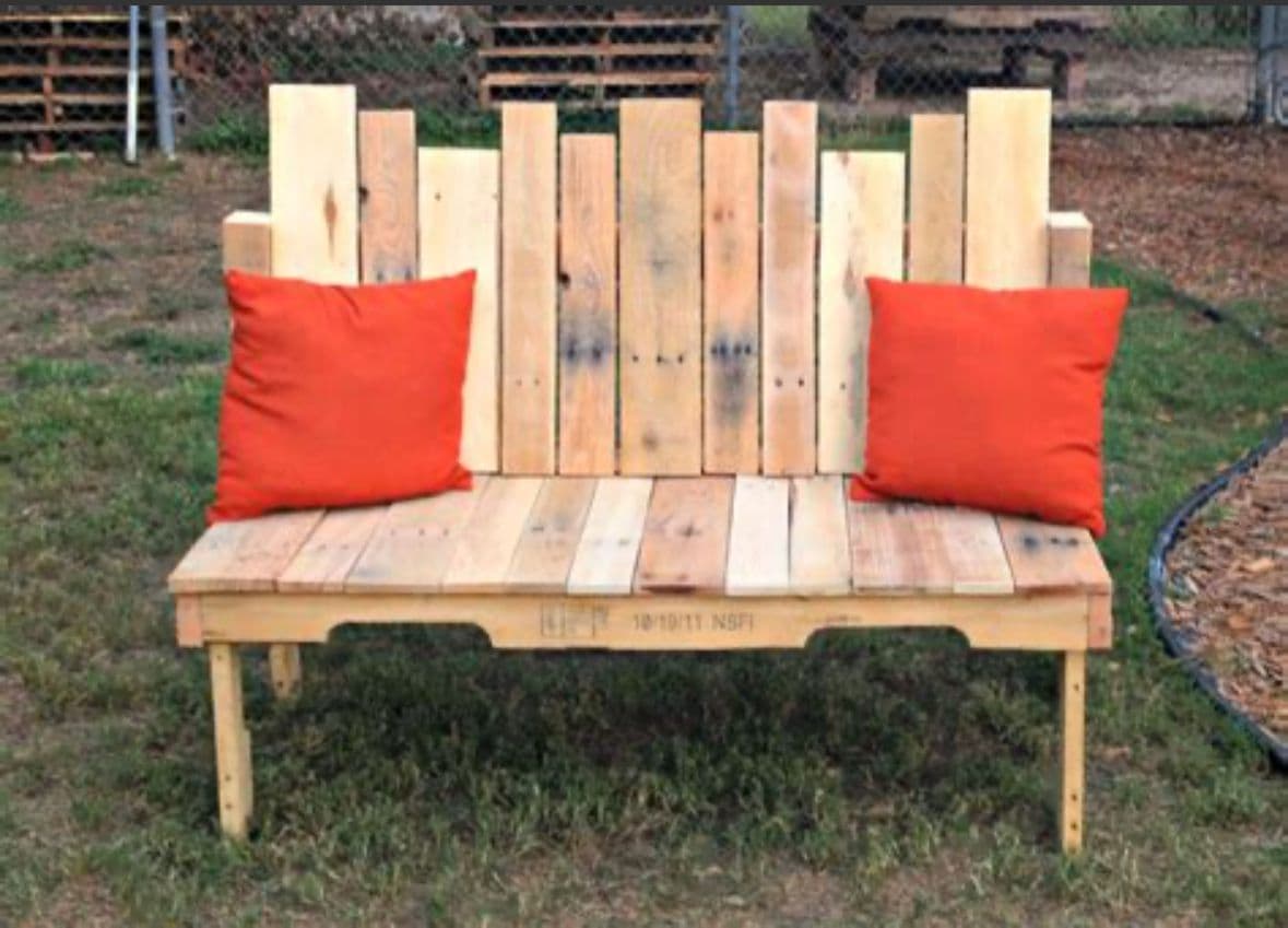 A rustic bench constructed from wooden pallet planks. The bench features a slatted seat and a backrest composed of vertically oriented planks of varying heights and widths, creating an uneven, staggered silhouette. It is accompanied by two square-shaped cushions in a vibrant orange hue, placed on the seat. — 3D model