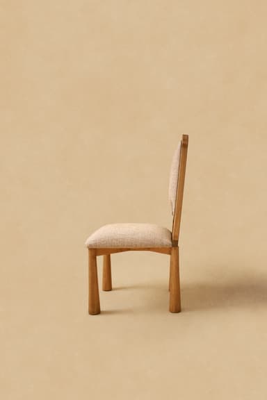 A modern armless chair featuring a wooden frame and upholstered seat and backrest. The chair's legs are stout and cylindrical, tapering slightly towards the floor. The backrest is gently curved, and both the seat and back are covered in a light beige, textured fabric. The wood has a natural, light tone, possibly oak or ash, with visible grain. — 3D model
