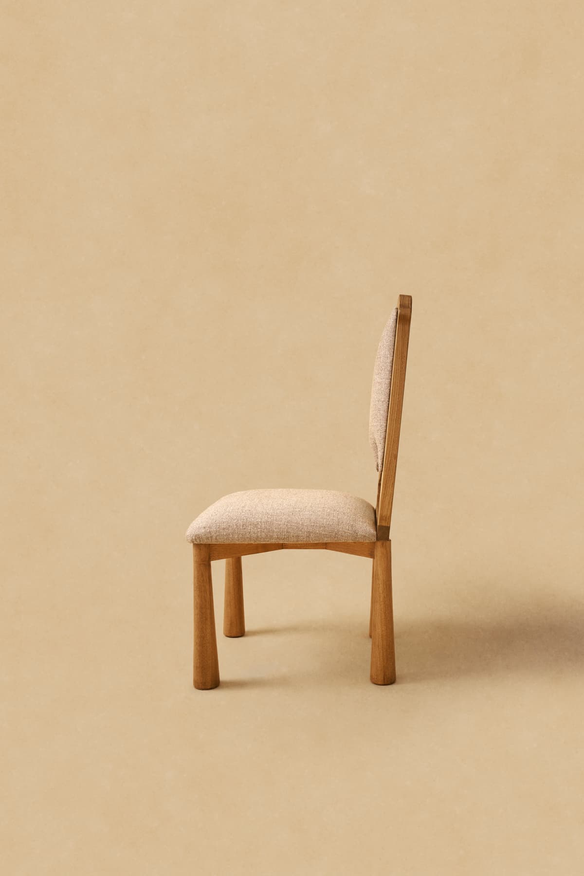 A modern armless chair featuring a wooden frame and upholstered seat and backrest. The chair's legs are stout and cylindrical, tapering slightly towards the floor. The backrest is gently curved, and both the seat and back are covered in a light beige, textured fabric. The wood has a natural, light tone, possibly oak or ash, with visible grain. — 3D model