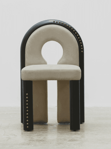A contemporary chair with a distinctive U-shaped backrest that forms a cutout. The seat and the curved portion of the backrest are upholstered in a light beige, suede-like fabric. The frame is composed of dark, possibly black or dark wood, elements. The outer curved frame of the backrest and the vertical supports of the legs are adorned with a series of small, brass or gold-colored studs. The legs are thick and rectangular, with a dark finish. The chair's design is modern and somewhat sculptural, featuring a negative space in the upper backrest. — 3D model