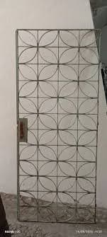 A tall, narrow, rectangular metal screen panel featuring a repeating geometric pattern of interconnected ovoid shapes. The panel is finished in a matte gray, suggesting a durable, industrial aesthetic. A small, rectangular reddish-brown handle is affixed to one vertical edge, indicating a functional aspect for opening, closing, or moving the panel. The design allows for partial visibility while offering a degree of privacy or decorative division. — 3D model