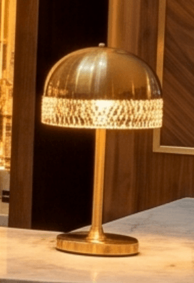 A contemporary table lamp with a brushed brass finish. The lamp features a domed shade with a textured band around its circumference, and a simple, slender stem rising from an oval base. A small finial is visible at the apex of the shade. — 3D model