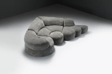 A large, multi-part sectional sofa featuring soft, organic, rounded forms, upholstered in a textured grey fabric. The sofa comprises several modules including corner pieces, armless central sections, and an end module that functions as an extended footrest or chaise. The overall design emphasizes comfort and a flowing, contemporary aesthetic. — 3D model