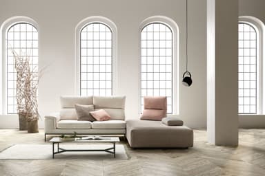 A modular sectional sofa with a left-arm facing chaise lounge. The sofa features a light-colored, textured fabric upholstery on the main seating sections and backrests. It includes adjustable headrests on the main sofa section and several throw pillows in varying shades of pink and grey. The chaise lounge section is upholstered in a taupe, textured fabric and has a low profile. The base of the sofa section is supported by slender black metal legs, creating a modern aesthetic. — 3D model