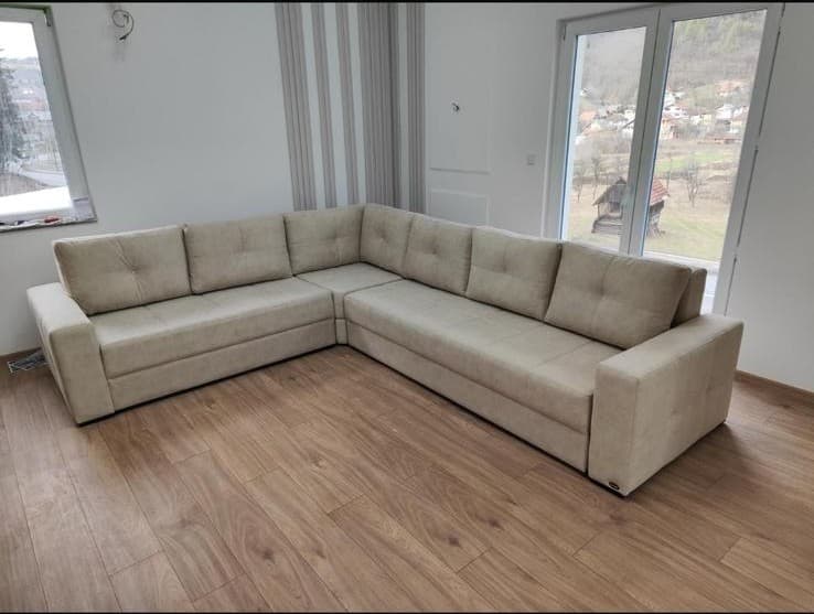 A large, modern L-shaped sectional sofa upholstered in a light beige or cream-colored fabric. The sofa features wide, flat track arms and back cushions with subtle tufting, providing a clean and contemporary aesthetic. It is positioned on a light wood laminate floor, suggesting a living room or lounge area, with large windows overlooking an outdoor scene. — 3D model