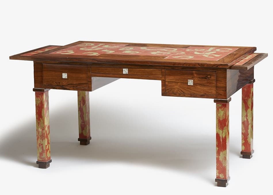 A finely crafted wooden desk with a rectangular top and four uniquely decorated legs. The desk features multiple drawers across its front, each fitted with a small, square metal pull. The top of the desk has a decorative inlay, primarily in red and lighter tones, with an intricate pattern. The legs are the most striking feature, exhibiting a marbled or painted effect in shades of red, yellow, and green, creating a distinctive, artistic appearance. — 3D model