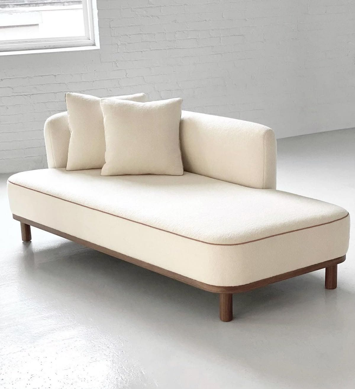 A modern cream bouclé upholstered chaise lounge with a single armrest and backrest, featuring contrasting piping and a dark wood base frame with cylindrical legs. Two matching throw pillows are included. — 3D model