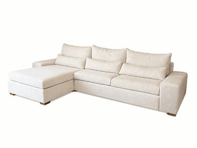 An L-shaped sectional sofa upholstered in a light beige, textured fabric. The sofa consists of three main seat cushions and a left-hand facing chaise. It features plush, loose back cushions and two cylindrical bolster pillows. The base is low-profile with short, square wooden block feet. — 3D model