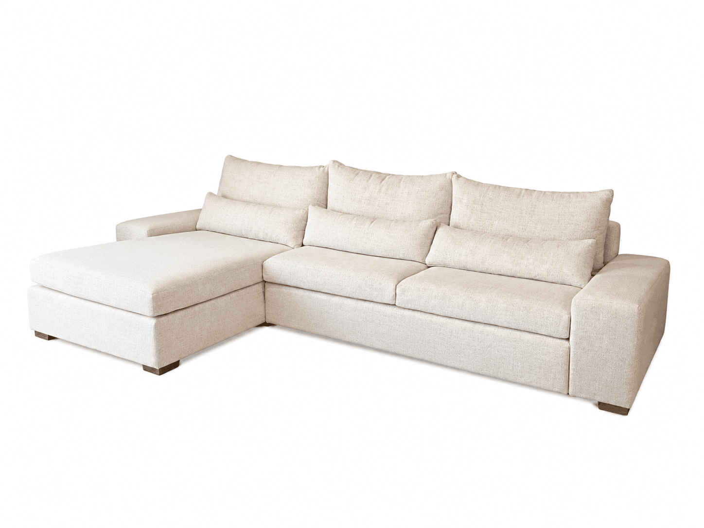 An L-shaped sectional sofa upholstered in a light beige, textured fabric. The sofa consists of three main seat cushions and a left-hand facing chaise. It features plush, loose back cushions and two cylindrical bolster pillows. The base is low-profile with short, square wooden block feet. — 3D model