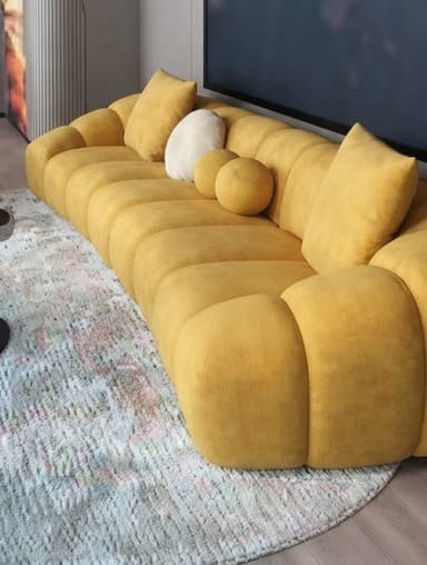 A long, contemporary sofa featuring a distinctive segmented design, with each section appearing deeply cushioned and rounded. It is upholstered in a textured, possibly bouclé fabric in a mustard yellow color. The low-profile design gives it a sculptural quality, and it is accompanied by a few square and round decorative pillows in matching and contrasting tones. — 3D model