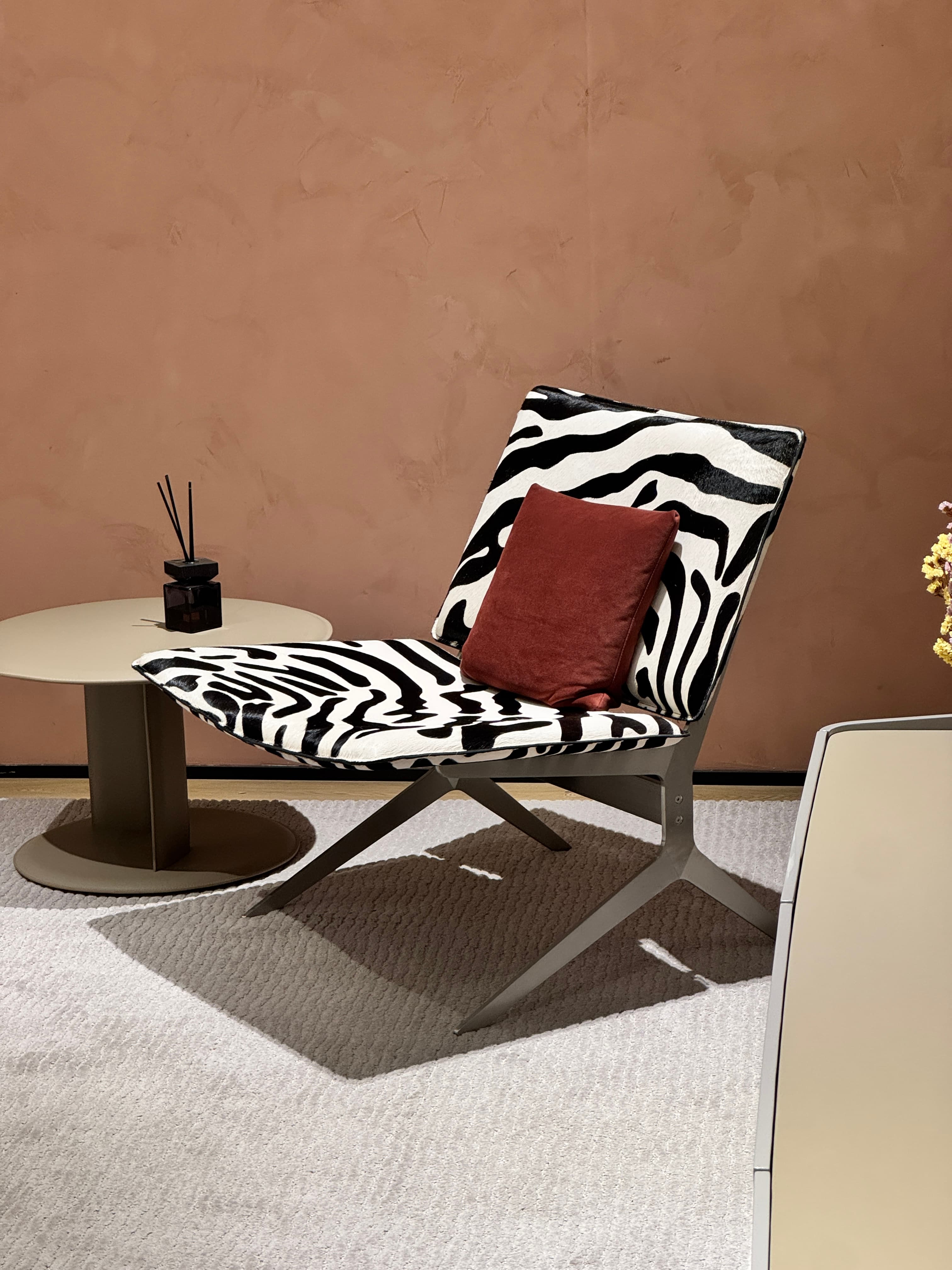 A modern lounge chair with a distinctive zebra print upholstery and a contrasting solid color throw pillow. The chair features a sleek, minimalist frame made of dark metal, with thin legs that splay outwards for stability. The seat and back are integrated, forming a continuous surface upholstered in a black and white zebra pattern. A small, square throw pillow in a reddish-brown hue is placed on the seat. — 3D model