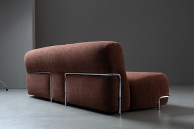 A compact, armless loveseat upholstered in a textured, dark reddish-brown fabric, featuring a minimalist design with a chrome-finished metal bar accent along the base. — 3D model