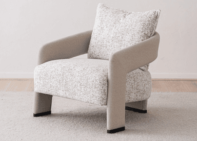 A contemporary upholstered armchair featuring a rounded silhouette and a neutral-toned, speckled fabric. The chair includes integrated, curved armrests and an upholstered backrest, complemented by a separate, plush back cushion with piped edges. The seat appears integrated, and the chair stands on short, upholstered legs or a low, concealed base, suitable for modern interior settings. — 3D model