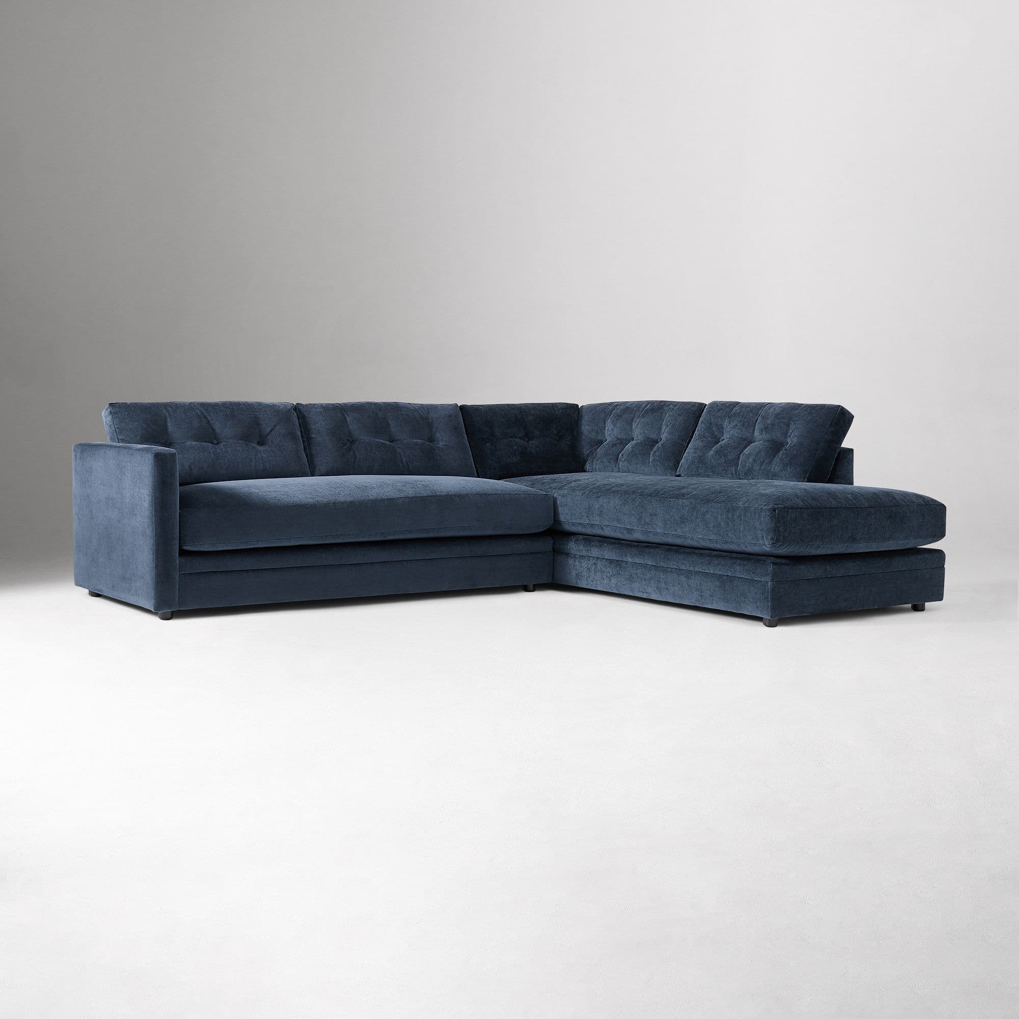 An L-shaped sectional sofa upholstered in a dark blue velvet fabric, featuring button-tufted back cushions and an integrated chaise lounge on the right-hand side. The sofa has a low profile with broad block arms and low, discreet feet. — 3D model