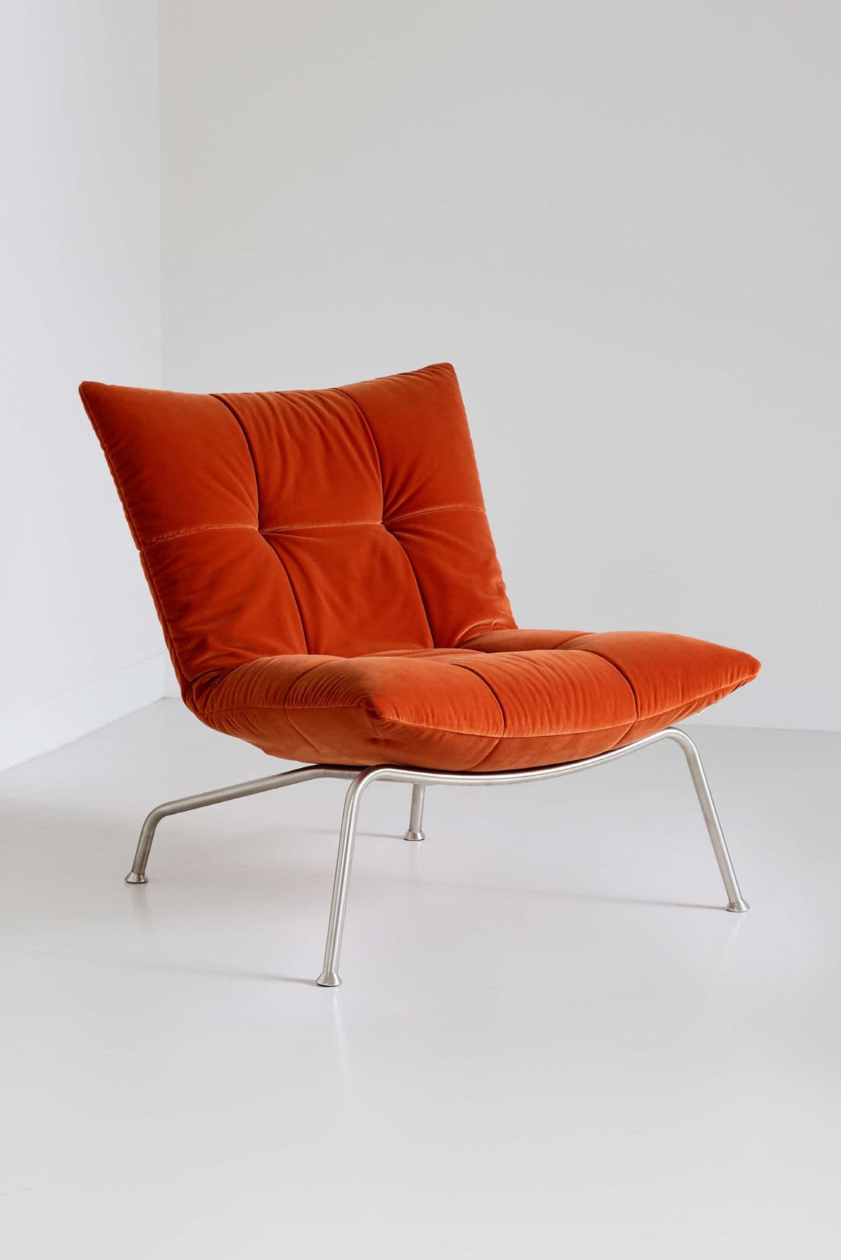 A modern lounge chair featuring plush, tufted burnt orange velvet upholstery and a sleek, minimalist tubular metal frame. The chair has a low profile, comprising a seat and backrest that appear deeply cushioned, forming a cohesive, inviting form. The metal frame supports the upholstered body with splayed legs, giving it a contemporary aesthetic. — 3D model
