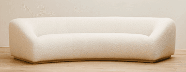 A contemporary, generously curved upholstered sofa in a light, neutral beige tone. The sofa features a continuous, rounded form with no visible legs, giving it a low-profile and substantial presence. The backrest and seat are seamlessly integrated, suggesting a cohesive, single-piece construction. The upholstery appears to be a textured fabric, possibly boucle or a fine-grained velvet, with subtle vertical seams segmenting the back and seat portions. Its design is minimalist, focusing on organic curves and comfort. — 3D model