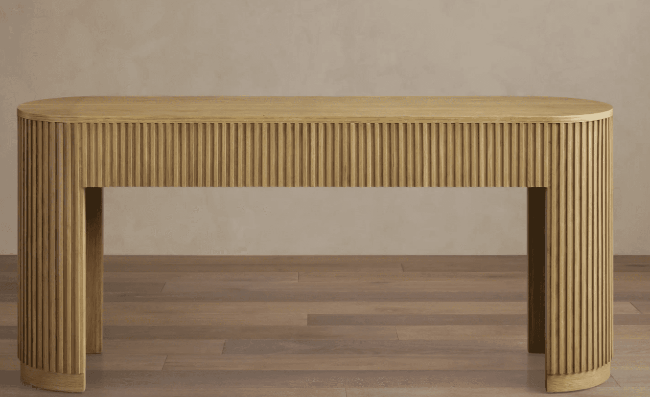 A narrow console table with a fluted design on its sides and front. It features a rounded rectangular top and two curved, solid legs that extend from the underside of the tabletop to the floor. The fluting runs vertically along the sides and the front panel, creating a textured appearance. The piece is constructed from wood and has a light, natural finish. — 3D model