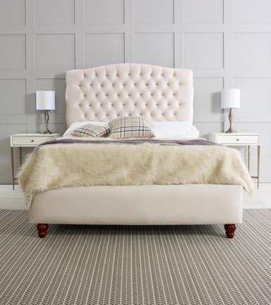 A cream-colored upholstered bed with a diamond-tufted headboard featuring crystal-like embellishments. The bed frame is also upholstered in a matching cream fabric. It is shown with a fluffy cream-colored throw blanket, two plaid throw pillows, and a gray knitted throw. The bed rests on four dark wooden carved legs. Two matching nightstands with drawers and brass accents are positioned on either side of the bed, each holding a white lampshade with a brass base. — 3D model