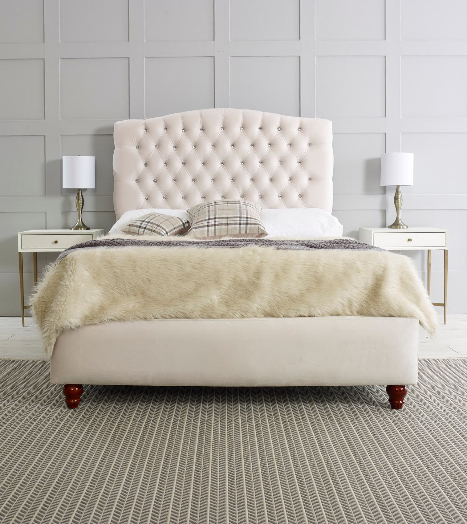 A cream-colored upholstered bed with a diamond-tufted headboard featuring crystal-like embellishments. The bed frame is also upholstered in a matching cream fabric. It is shown with a fluffy cream-colored throw blanket, two plaid throw pillows, and a gray knitted throw. The bed rests on four dark wooden carved legs. Two matching nightstands with drawers and brass accents are positioned on either side of the bed, each holding a white lampshade with a brass base. — 3D model