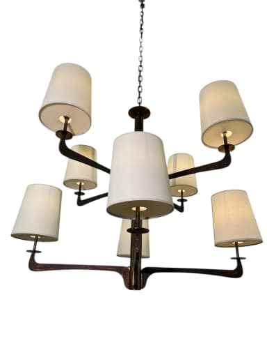A modern multi-arm chandelier featuring a dark bronze-finished metal frame and eight tapered, drum-style fabric shades. The central column extends from a chain suspension, with eight arms branching out, each supporting an upward-facing light socket covered by a cream-colored textile shade. The metalwork exhibits a rustic or aged patina, suggesting a blend of modern design with traditional material treatment. — 3D model