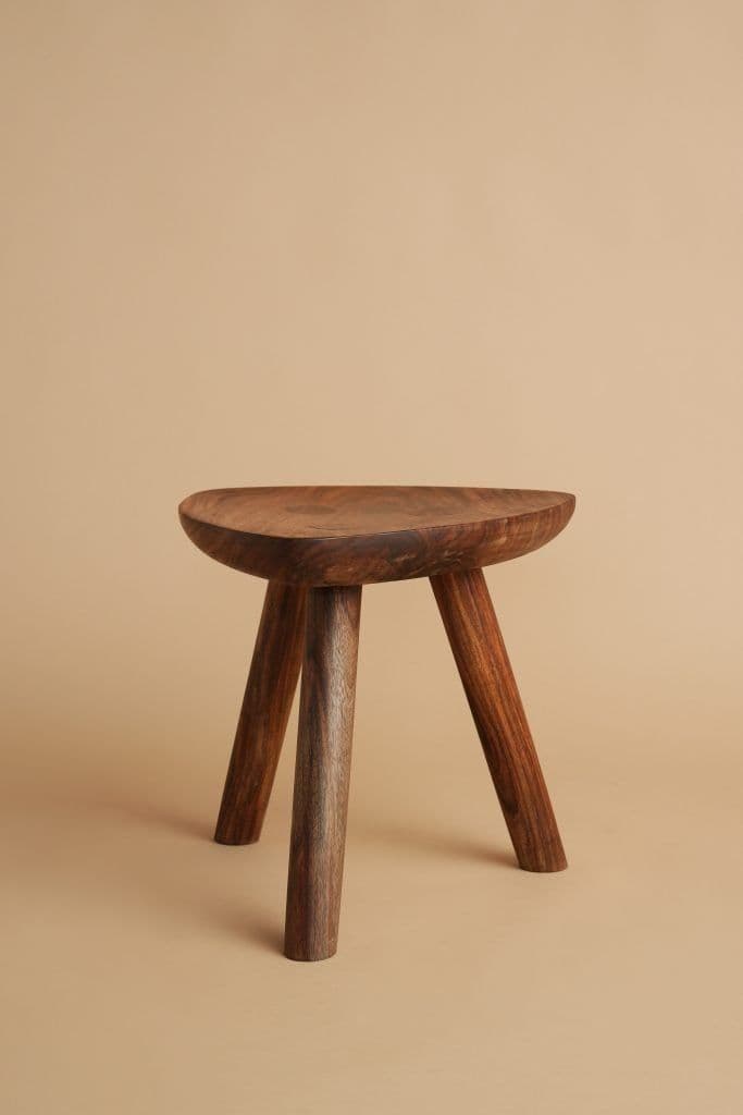 A sculptural three-legged stool crafted from solid dark wood, featuring a concave, saddle-shaped seat and three splayed, tapered legs. The design emphasizes natural forms and smooth, rounded edges. — 3D model