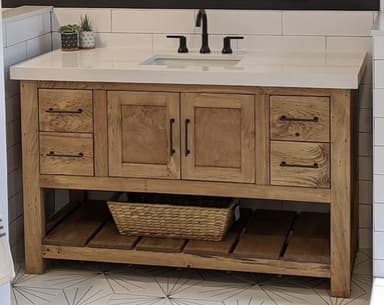 A rustic-style bathroom vanity constructed from light-toned oak wood, featuring a white engineered stone countertop with an integrated rectangular ceramic sink. The vanity includes two functional drawers and a two-door cabinet in the center, all equipped with matte black bar pull handles. Below the main cabinet, there is an open slatted shelf for additional storage. A matte black gooseneck faucet and matching soap dispenser are mounted on the countertop. The overall design emphasizes clean lines and natural materials, suitable for a contemporary or farmhouse aesthetic. — 3D model