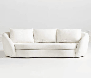 A close-up view of a cream-colored sofa, focusing on the armrest and seat cushions. The upholstery appears to be a textured boucle or similar woven fabric. The armrest has a rounded, curved profile, and the seat cushions are plush with visible stitching. The base of the sofa, where it meets the floor, is partially visible and appears to be a dark wood. — 3D model