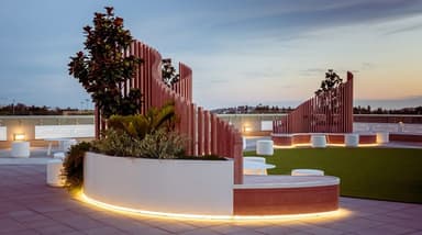 A modern, modular curved outdoor seating system featuring light-toned wood construction with white upholstered cushions and white solid-surface sections. The design incorporates integrated planters filled with greenery and a slatted wooden backrest that varies in height. Subtly lit accent lighting is visible along the base of the white sections. Two small white cylindrical stools and a small white round table are positioned within the curve of the bench. — 3D model