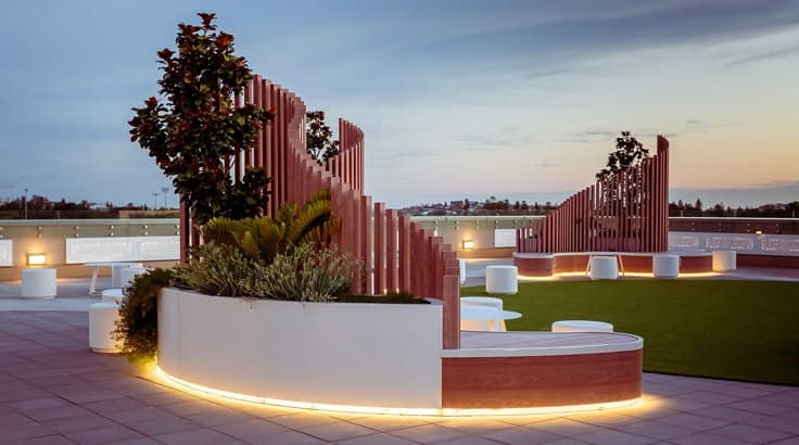 A modern, modular curved outdoor seating system featuring light-toned wood construction with white upholstered cushions and white solid-surface sections. The design incorporates integrated planters filled with greenery and a slatted wooden backrest that varies in height. Subtly lit accent lighting is visible along the base of the white sections. Two small white cylindrical stools and a small white round table are positioned within the curve of the bench. — 3D model