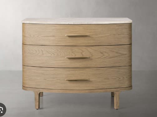 A chest of drawers with a rounded front profile, featuring three drawers stacked vertically. The top surface appears to be made of a light-colored stone, possibly marble or quartz. The main body and drawer fronts are constructed from light-colored wood with a visible grain pattern. Each drawer is equipped with a horizontal, metallic handle. The unit stands on four tapered legs that angle slightly outwards. — 3D model